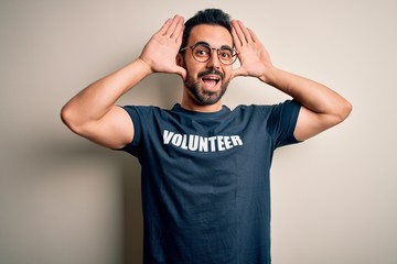 Handsome man with beard wearing t-shirt with volunteer message over white background Smiling cheerful playing peek a boo with hands showing face. Surprised and exited
