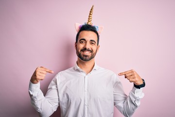 Young handsome man with beard wearing funny unicorn diadem over pink background looking confident with smile on face, pointing oneself with fingers proud and happy. © Krakenimages.com