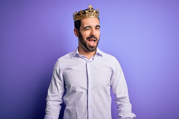 Young handsome man with beard wearing golden crown of king over purple background winking looking...