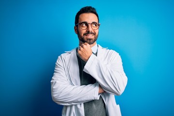Young handsome doctor man with beard wearing coat and glasses over blue background with hand on chin thinking about question, pensive expression. Smiling with thoughtful face. Doubt concept.