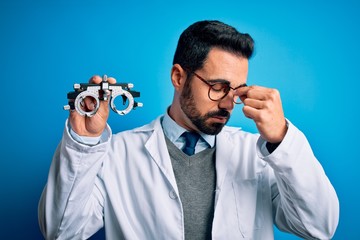 Young handsome optical man with beard holding optometry glasses over blue background tired rubbing...