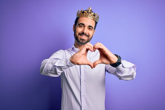 Young handsome man with beard wearing golden crown of king over purple background smiling in love doing heart symbol shape with hands. Romantic concept.