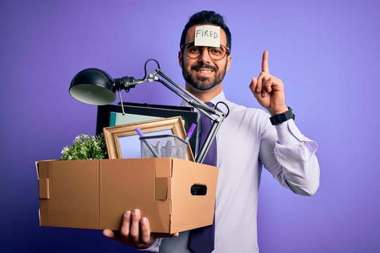 Young handsome businessman with beard holding cardboard box with fired reminder on head surprised with an idea or question pointing finger with happy face, number one