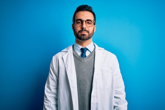 Young handsome doctor man with beard wearing coat and glasses over blue background Relaxed with serious expression on face. Simple and natural looking at the camera.