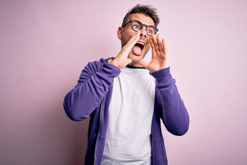 Young handsome man wearing purple sweatshirt and glasses standing over pink background Shouting angry out loud with hands over mouth