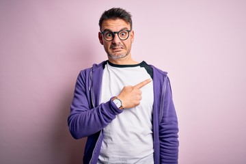 Young handsome man wearing purple sweatshirt and glasses standing over pink background Pointing...