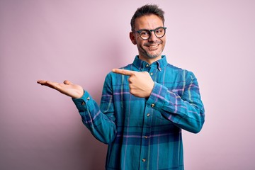 Young handsome man wearing casual shirt and glasses standing over isolated pink background amazed and smiling to the camera while presenting with hand and pointing with finger.