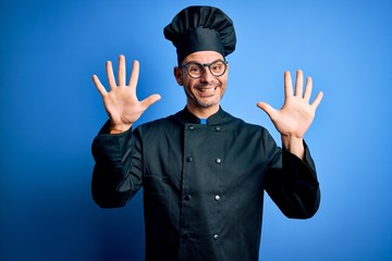 Young handsome chef man wearing cooker uniform and hat over isolated blue background showing and pointing up with fingers number ten while smiling confident and happy.