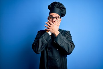 Young handsome chef man wearing cooker uniform and hat over isolated blue background shocked covering mouth with hands for mistake. Secret concept.