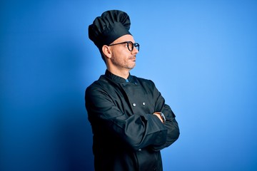 Young handsome chef man wearing cooker uniform and hat over isolated blue background looking to the side with arms crossed convinced and confident