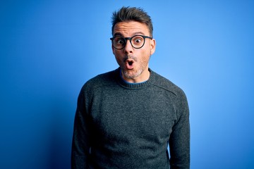 Young handsome man wearing casual sweater and glasses standing over blue background afraid and shocked with surprise expression, fear and excited face.