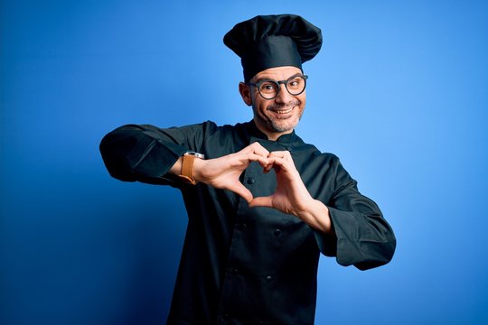 Young handsome chef man wearing cooker uniform and hat over isolated blue background smiling in love doing heart symbol shape with hands. Romantic concept.