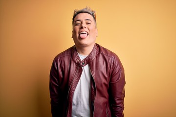 Young handsome modern man wearing fashion leather jacket and sunglasses over yellow background sticking tongue out happy with funny expression. Emotion concept.