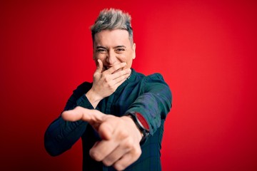 Young handsome modern man wearing elegant green shirt over red isolated background laughing at you, pointing finger to the camera with hand over mouth, shame expression