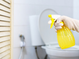 Close up hand with glove holding yellow spray bottle of cleanser over blurry white toilet background.