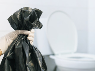 Close up hand with glove holding black garbage bag over blurry white toilet background.