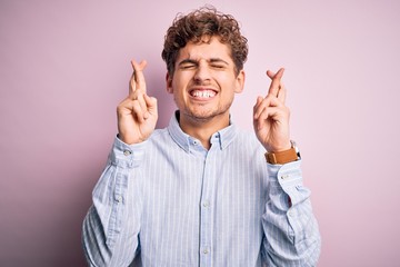 Young blond handsome man with curly hair wearing striped shirt over white background gesturing finger crossed smiling with hope and eyes closed. Luck and superstitious concept.