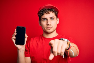 Young blond delivery man with curly hair wearing cap holding smartphone over red background pointing with finger to the camera and to you, hand sign, positive and confident gesture from the front