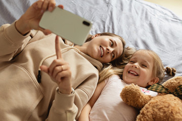 Mom and daughter watching video on smartphone with smiley face alone on the bed, child using mobile phone with happy face at home. Stay at home quarantine coronavirus COVID-19 pandemic prevention.