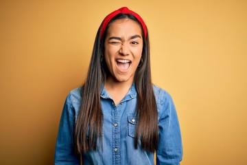 Young beautiful asian woman wearing casual denim shirt and diadem over yellow background winking...