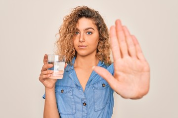 Young beautiful blonde woman with blue eyes drinking glass of water over white background with open hand doing stop sign with serious and confident expression, defense gesture