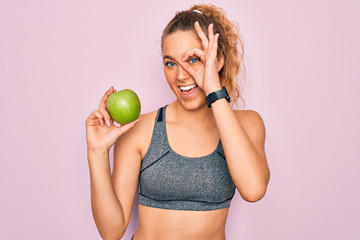 Young beautiful blonde sporty woman with blue eyes holding healthy green apple fruit with happy face smiling doing ok sign with hand on eye looking through fingers
