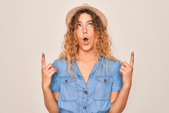 Young beautiful woman with blue eyes wearing casual denim dress and summer hat amazed and surprised looking up and pointing with fingers and raised arms.