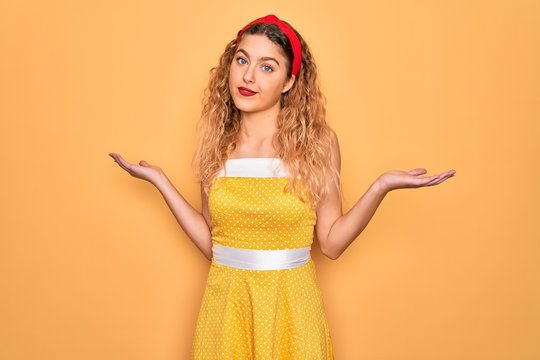 Beautiful blonde pin-up woman with blue eyes wearing diadem standing over yellow background clueless and confused expression with arms and hands raised. Doubt concept.