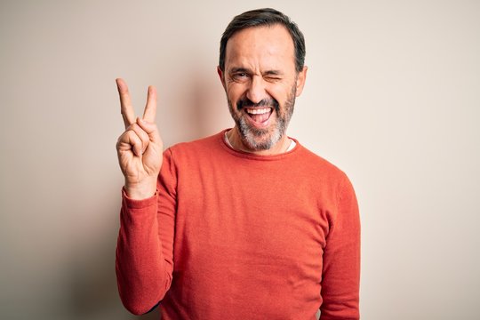 Middle age hoary man wearing casual orange sweater standing over isolated white background smiling with happy face winking at the camera doing victory sign with fingers. Number two.