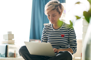 Beautiful Asian woman sitting on bed & working from home on laptop