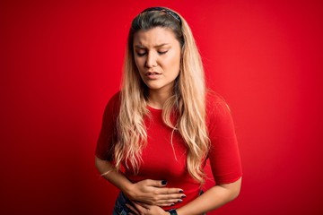 Young beautiful blonde woman wearing casual t-shirt standing over isolated red background with hand on stomach because indigestion, painful illness feeling unwell. Ache concept.