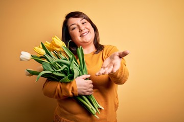 Beautiful plus size woman holding romantic bouquet of natural tulips flowers over yellow background smiling cheerful offering palm hand giving assistance and acceptance.