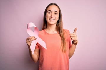 Young beautiful woman holding pink cancer ribbon standing over isolated pink background happy with big smile doing ok sign, thumb up with fingers, excellent sign