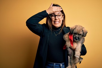 Young beautiful brunette woman holding cute puppy pet over isolated yellow background stressed with hand on head, shocked with shame and surprise face, angry and frustrated. Fear and upset for mistake