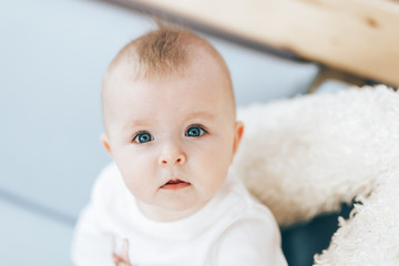 little very beautiful baby girl, looking at camera with beautiful blue eyes, cute baby sitting at home, light color interior.