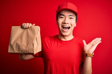 Young handsome chinese delivery man holding takeaway paper bag with food very happy and excited, winner expression celebrating victory screaming with big smile and raised hands