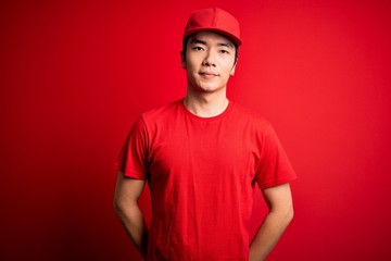 Young handsome chinese delivery man wearing cap standing over isolated red background with serious expression on face. Simple and natural looking at the camera.