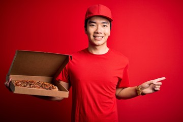 Young handsome chinese delivery man holding deliver box with delicious Italian pizza very happy pointing with hand and finger to the side