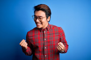 Naklejka premium Young handsome chinese man wearing casual shirt and glasses over blue background very happy and excited doing winner gesture with arms raised, smiling and screaming for success. Celebration concept.