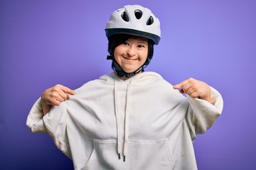 Young down syndrome cyclist woman wearing security bike helmet over purple background looking confident with smile on face, pointing oneself with fingers proud and happy.