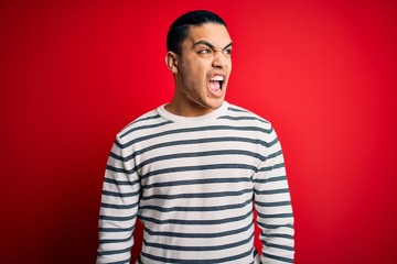 Obraz premium Young brazilian man wearing casual striped t-shirt standing over isolated red background angry and mad screaming frustrated and furious, shouting with anger. Rage and aggressive concept.