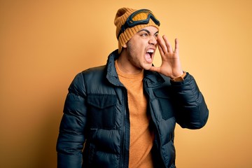Young brazilian skier man wearing snow sportswear and ski goggles over yellow background shouting and screaming loud to side with hand on mouth. Communication concept.