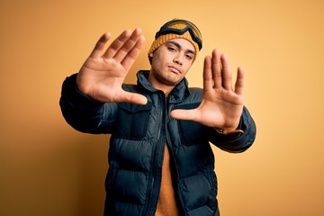 Young brazilian skier man wearing snow sportswear and ski goggles over yellow background doing frame using hands palms and fingers, camera perspective