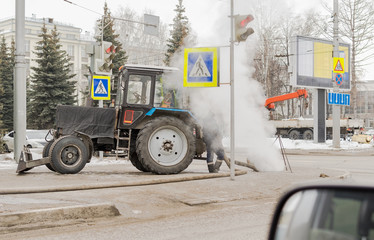a tractor of the municipal social service stands on a city street against the background of passing cars, a crane and thick smoke from the manhole of a faulty sewer