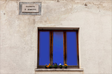 Finestra con tende blù in piazza Zorutti con muro bianco e fiori alla finestra. Cividale Del Friuli.