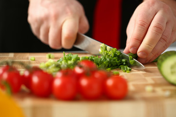 Cook holds knife in hand and cuts on cutting board green onions for salad or fresh vegetable soup with vitamins. Raw food and vegetarian recipe book in modern society popular concept.