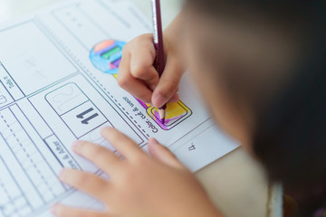 An Asian kindergarten girl is doing her homework with intentional coloring and beautifully drained on the homework sheet, selective focus on color pencil.