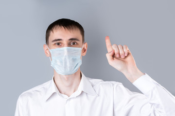 Man in a medical mask. A handsome dark-haired man in a white medical uniform, shirt and medical mask  looking at camera and shows a thumbs-up gesture on a gray background. Chinese Coronavirus 