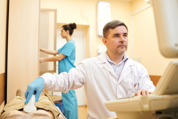 Serious mature doctor in lab coat doing ultrasound scanning while examining abdomen of patient