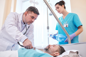 Fototapeta premium Concentrated mature doctor using stethoscope while examining lungs of patient in hospital room, nurse with ambu mask standing behind bed